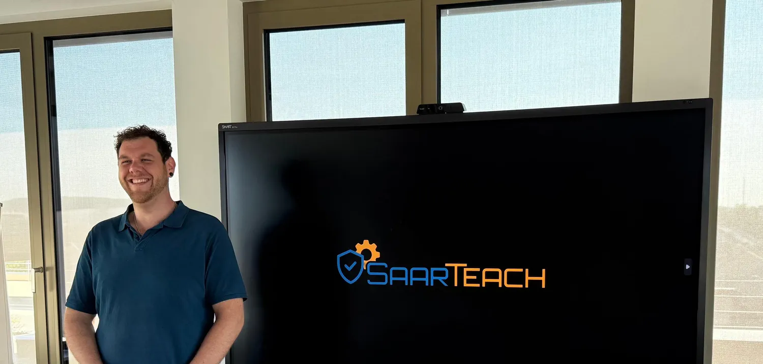 SaarTeach - Dozent für elektrotechnik und arbeitschutz - Thorsten Zimmer steht neben einem großen Bildschirm mit dem „SAARTEACH“-Logo.